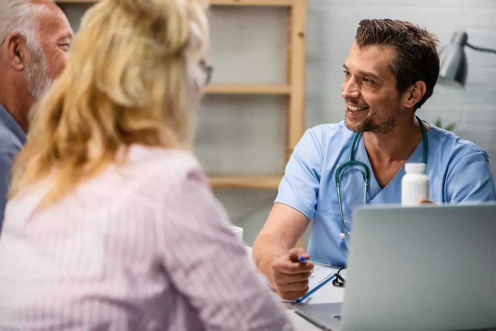  A smiling nurse or doctor in blue scrubs consults with a mature couple, likely discussing a treatment plan or progress in spinal fusion rehabilitation.