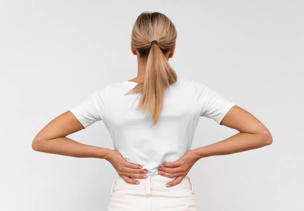 A woman in casual clothing standing with her back to the camera, places her hands on her lower back, indicating discomfort or pain possibly associated with spinal fusion rehabilitation exercises.