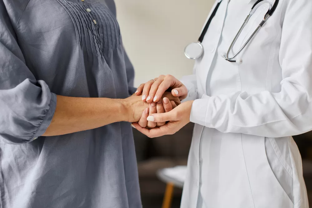 A healthcare professional in a white coat and stethoscope holds the hands of an elderly patient in a comforting gesture, symbolizing support during spinal fusion rehabilitation.